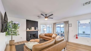 A modern living room with a leather sofa, a fireplace, a ceiling fan, and large windows. - Entertainment Galore in 4BR Oasis w/ Pool! (North Las Vegas)