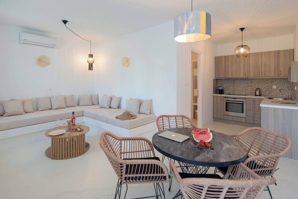 A modern living room with a sofa, round dining table, wicker chairs, and a kitchen area with wooden cabinets and a sink.