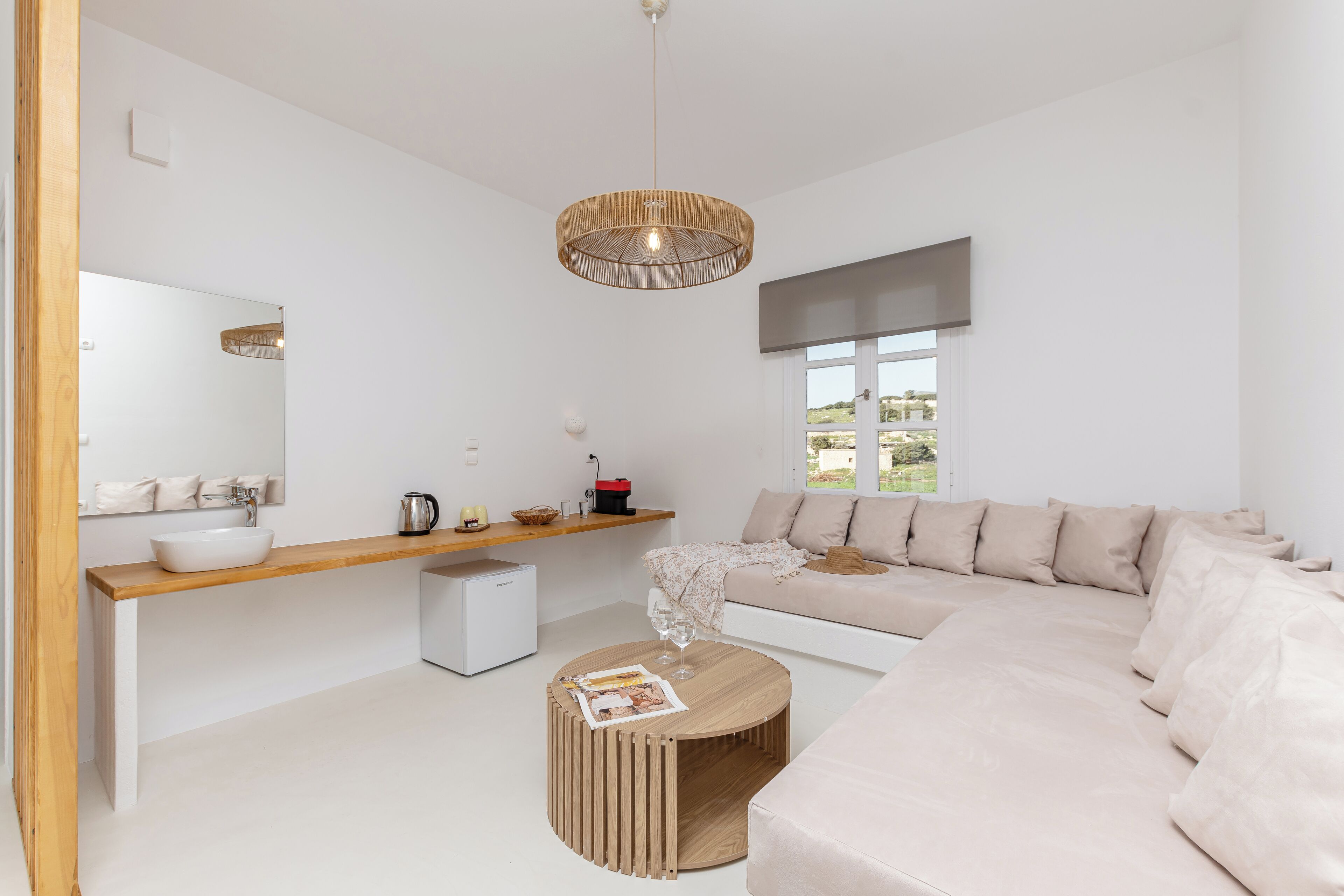 A modern living room with a large beige sofa, a wooden coffee table, a round wooden side table, a white sink, and a large window with a view of greenery.