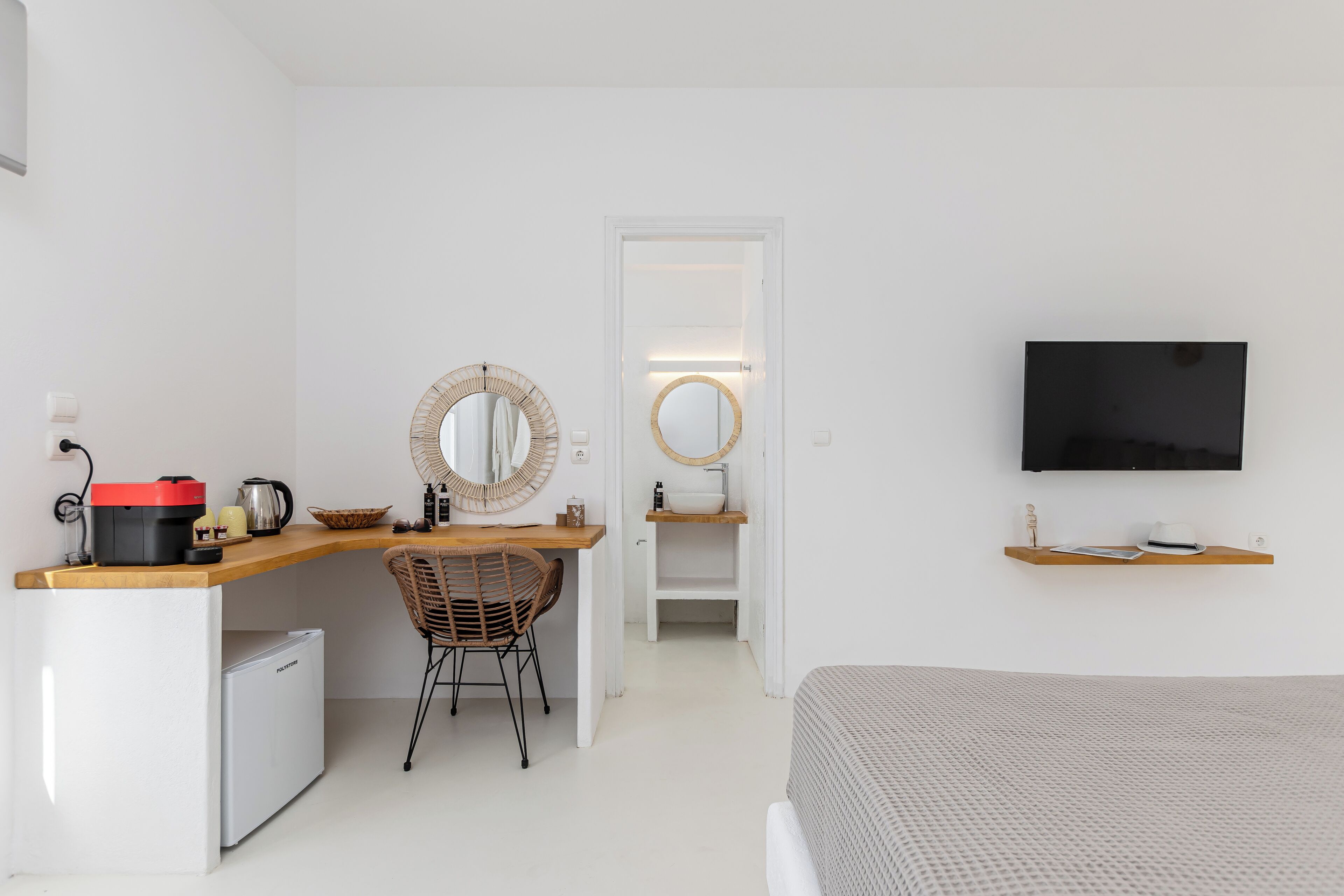 A bedroom with a bed, a desk with a chair, a small refrigerator, a television mounted on the wall, and a bathroom visible through an open door.