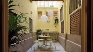 Un patio estrecho con una mesa y sillas, plantas en macetas y farolillos de papel colgantes.