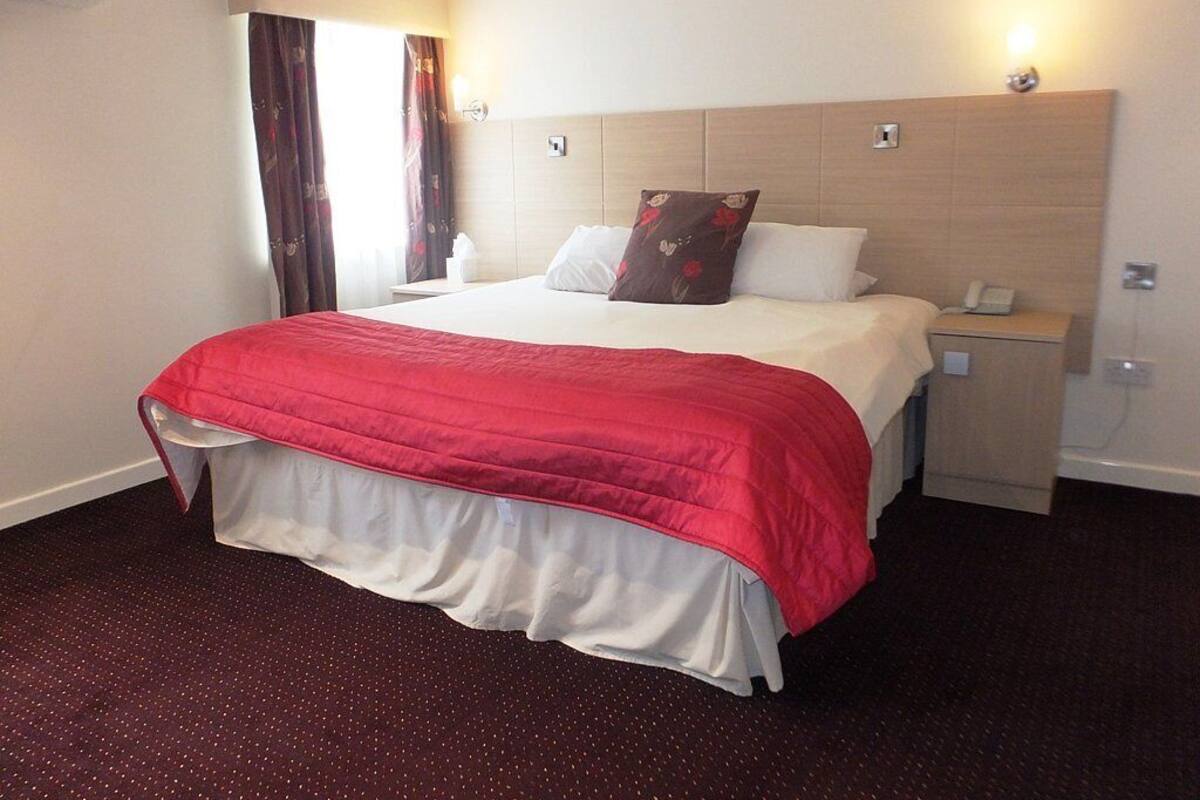 Ein Hotelzimmer mit einem großen Bett, einem Nachttisch mit Telefon und einem Fenster mit Vorhängen.