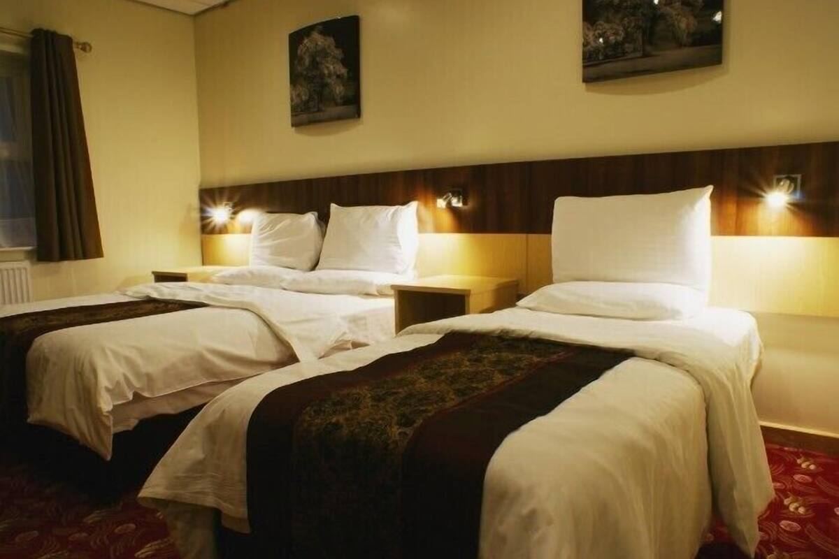 Ein Hotelzimmer mit zwei Betten, jeweils mit weißer Bettwäsche und einer dunkelbraunen Tagesdecke. An beiden Seiten der Betten befinden sich Wandlampen, und über dem linken Bett hängt ein Gemälde.