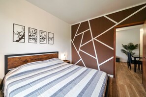 A bedroom with a bed, a wall with geometric patterns, and a dining area visible through an open door.