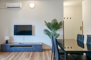 A modern living room with a flat-screen TV mounted on the wall, a dark wooden entertainment unit, a dining table with black chairs, and a potted plant.