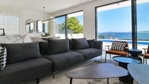 Una sala de estar moderna con un sofá seccional gris, una mesa de centro redonda y vista al mar a través de ventanas amplias.