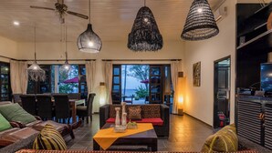 Un vaste espace de vie comprenant un canapé, des fauteuils et une table basse. On y trouve trois luminaires suspendus et un ventilateur de plafond. La pièce est dotée de grandes fenêtres avec des rideaux et offre une vue sur l’extérieur.