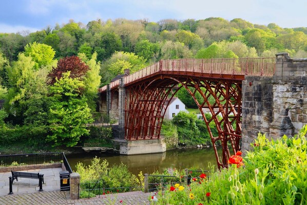 Un puente de piedra histórico con estructura metálica, un edificio blanco junto al río y un banco con vistas a la escena.