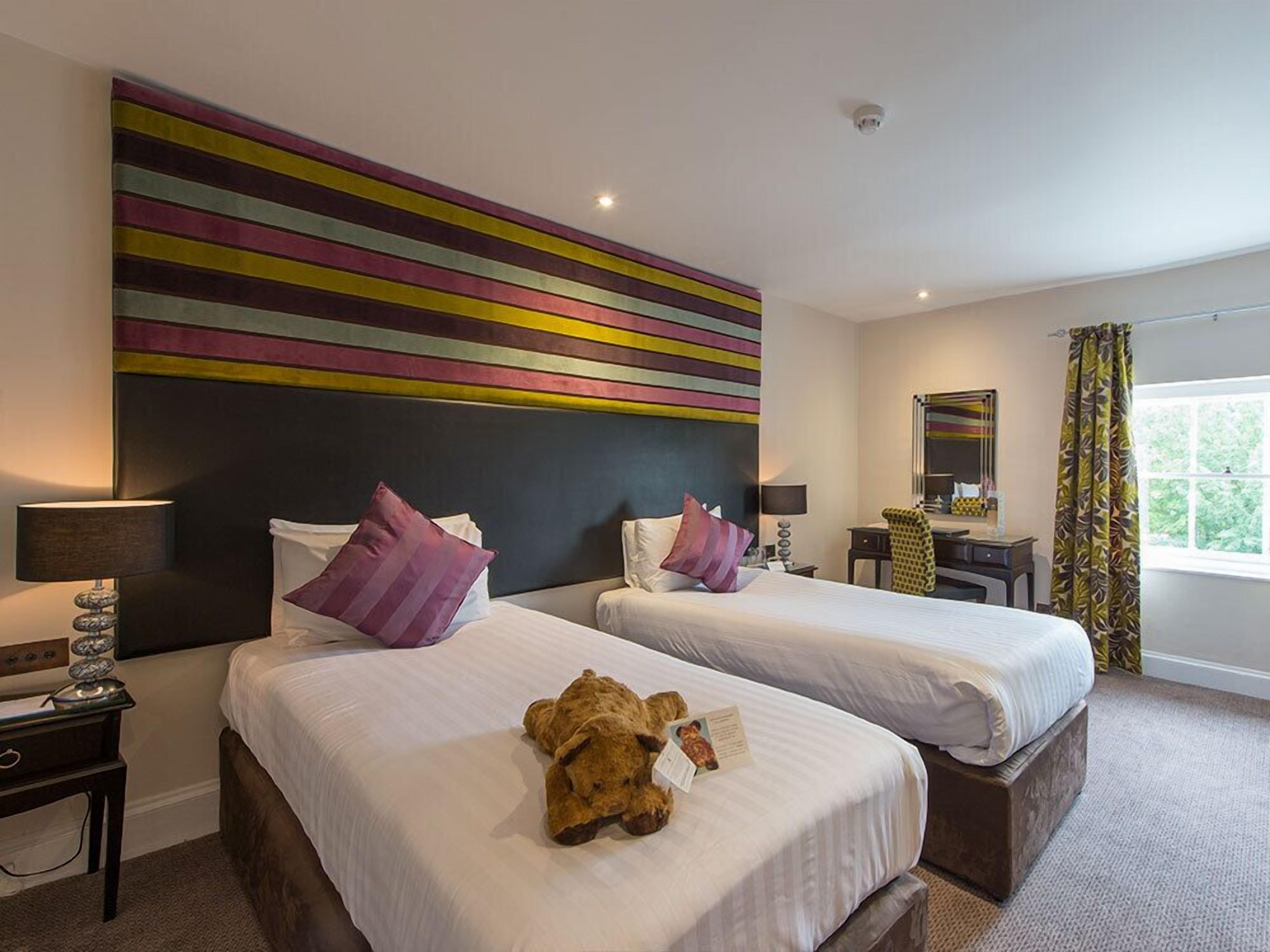 A hotel room with two beds, a large abstract painting on the wall, a desk with a chair, a mirror, and a window with curtains.