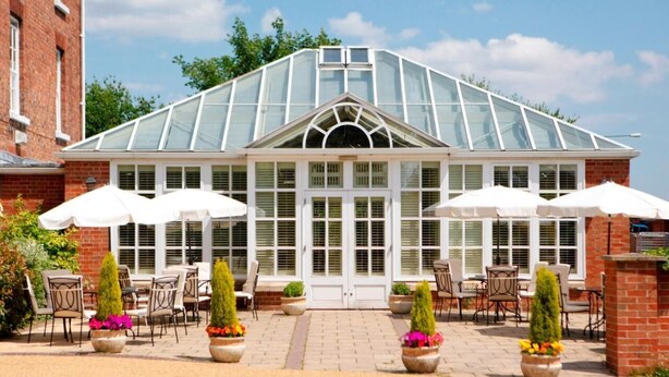 A greenhouse-style building with a glass roof, surrounded by outdoor seating with white umbrellas, and potted plants. - Hadley Park House Hotel (Telford)