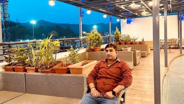 A man seated on a wicker chair in an outdoor seating area with potted plants and a view of the mountains. - Hotel City Pride (Haridwar)