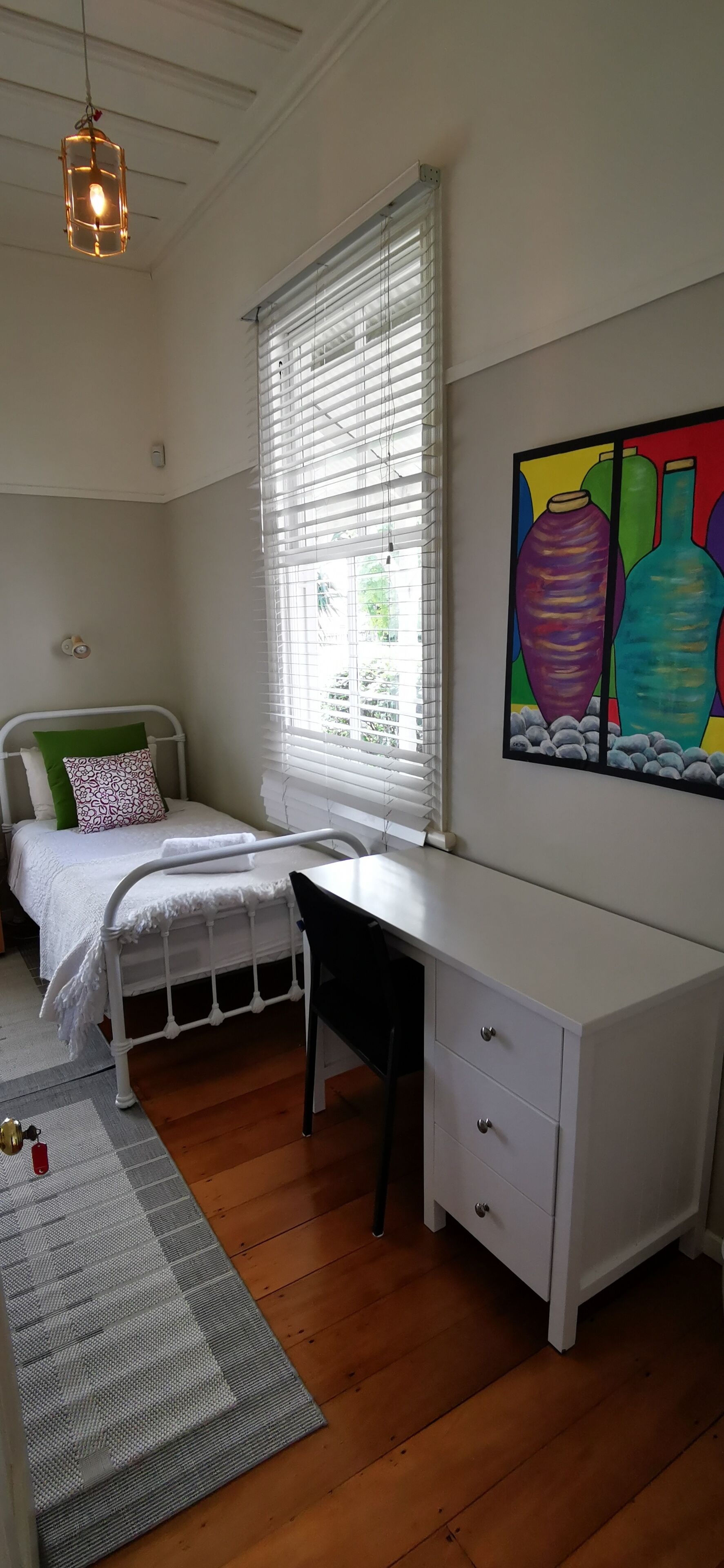 A bedroom with a bed, a desk, a chair, a radiator, and a window with blinds.