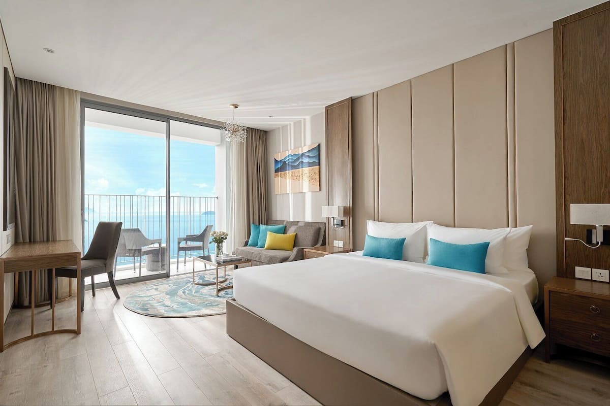 A hotel room with a large bed, a desk, a chair, a small table, and a view of the sea.
