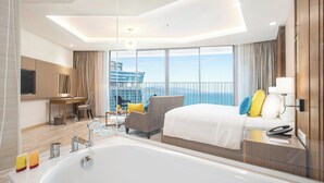 A modern hotel room with a large bed, a sitting area with a chair and table, a bathtub, and a view of the cityscape.