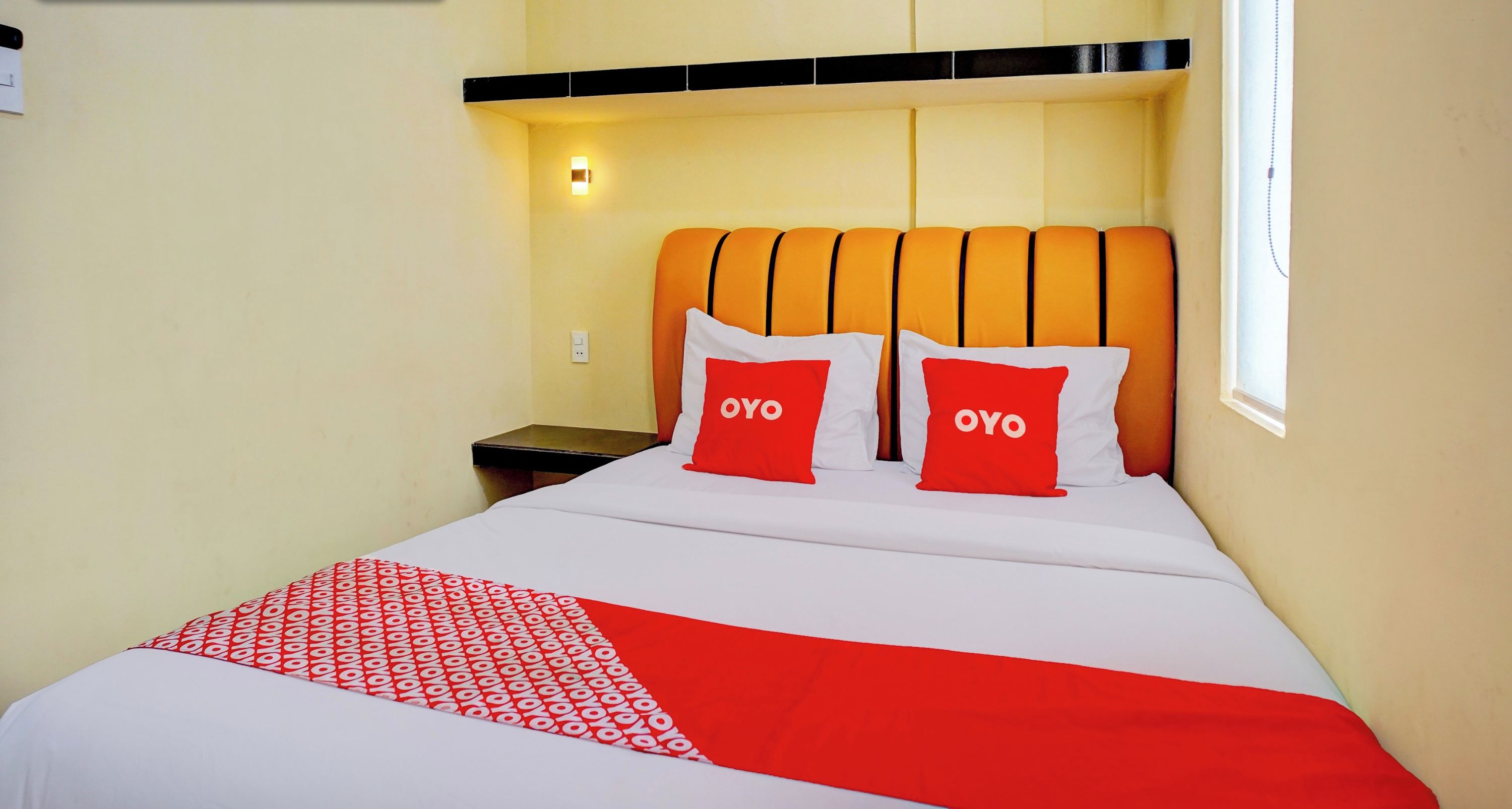 Habitación de hotel con una cama, dos almohadas rojas con el texto "OYO", una mesita de noche y un cartel en la pared que dice "GESTIONADO POR OYO".