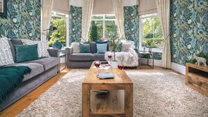 A cozy living room with a grey sofa, a wooden coffee table, and a white furry chair. There are two windows with curtains, and a plant on the windowsill. - Rosemount Lakes Townhouse with Hot Tub | Ridgestone (Windermere)