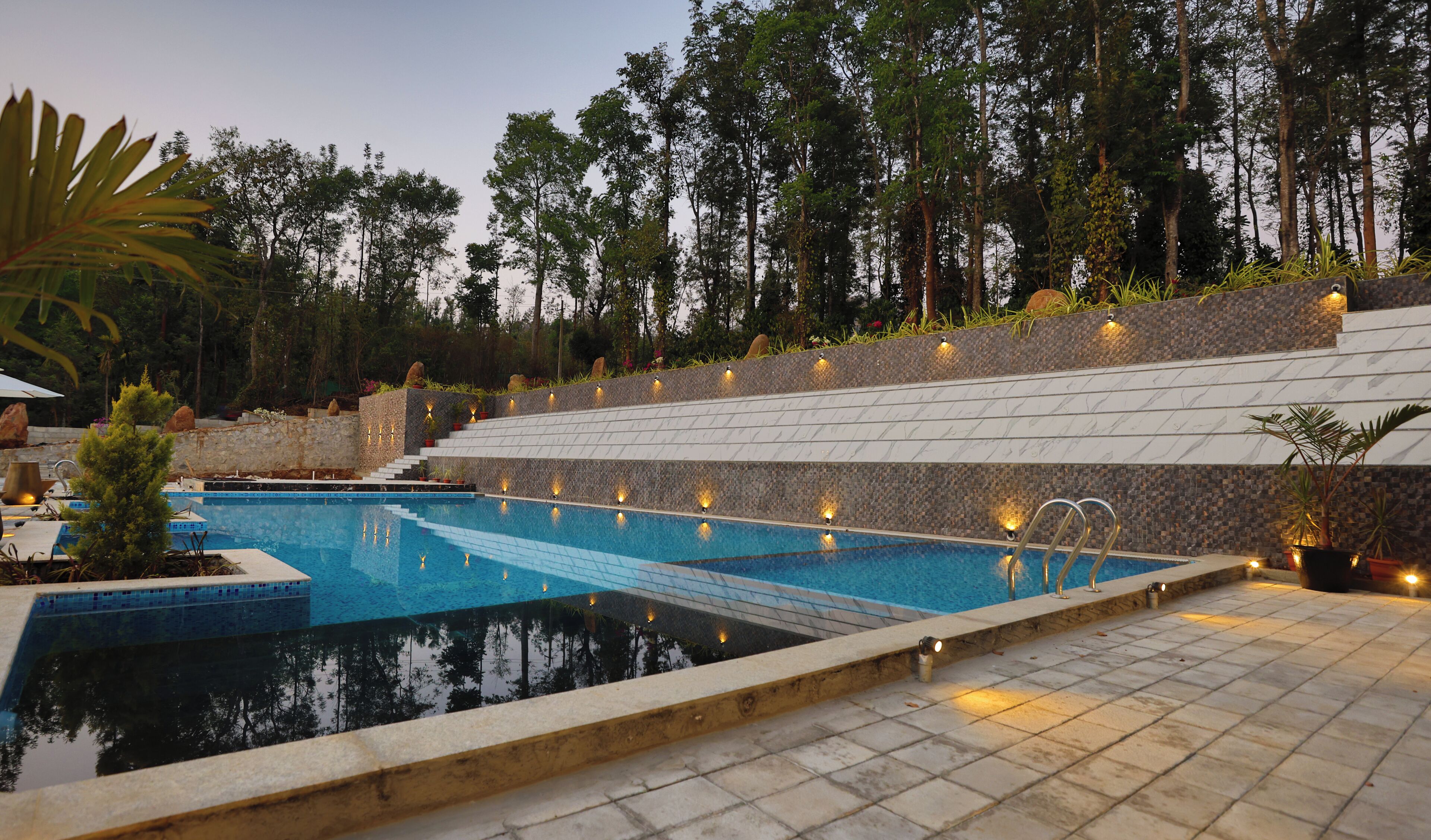A swimming pool with tiered seating and ambient lighting, surrounded by trees and a stone wall.