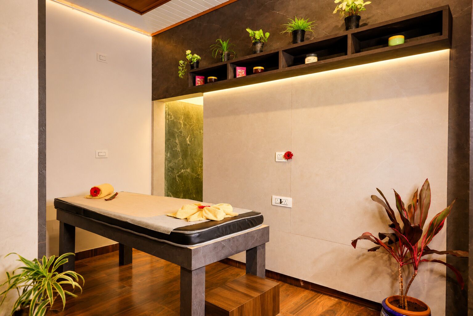 A massage table with towels and a flower, a shelf with plants, and a potted plant on the floor.