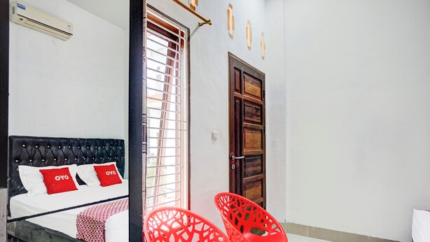 A bedroom with a bed, a window with blinds, a wooden door, and two red plastic chairs.