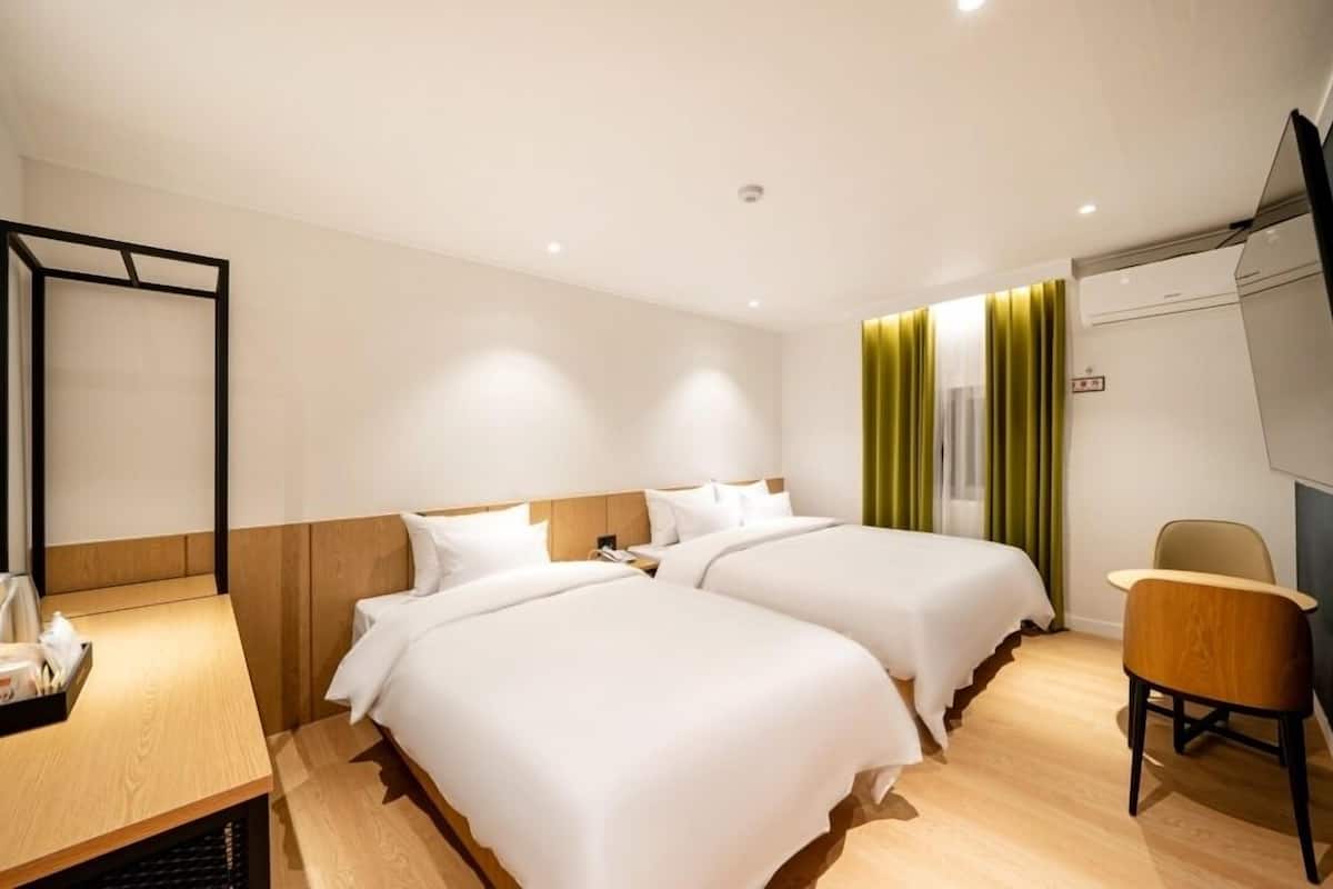Ein Hotelzimmer mit zwei Betten, einem Schreibtisch mit Stuhl, einem Fernseher und einem Fenster mit Vorhängen.
