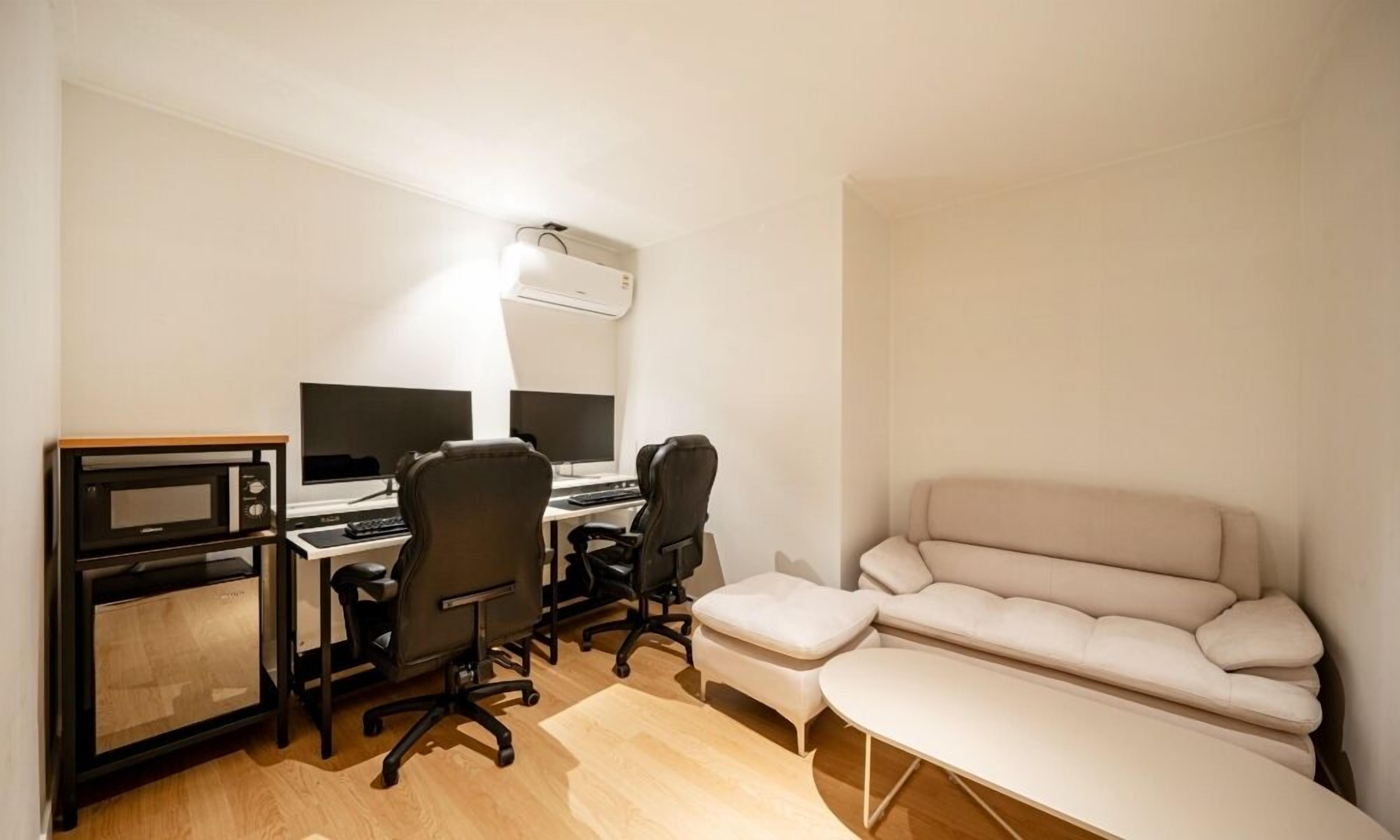 A compact room with a desk, two monitors, a chair, a sofa, and a microwave.