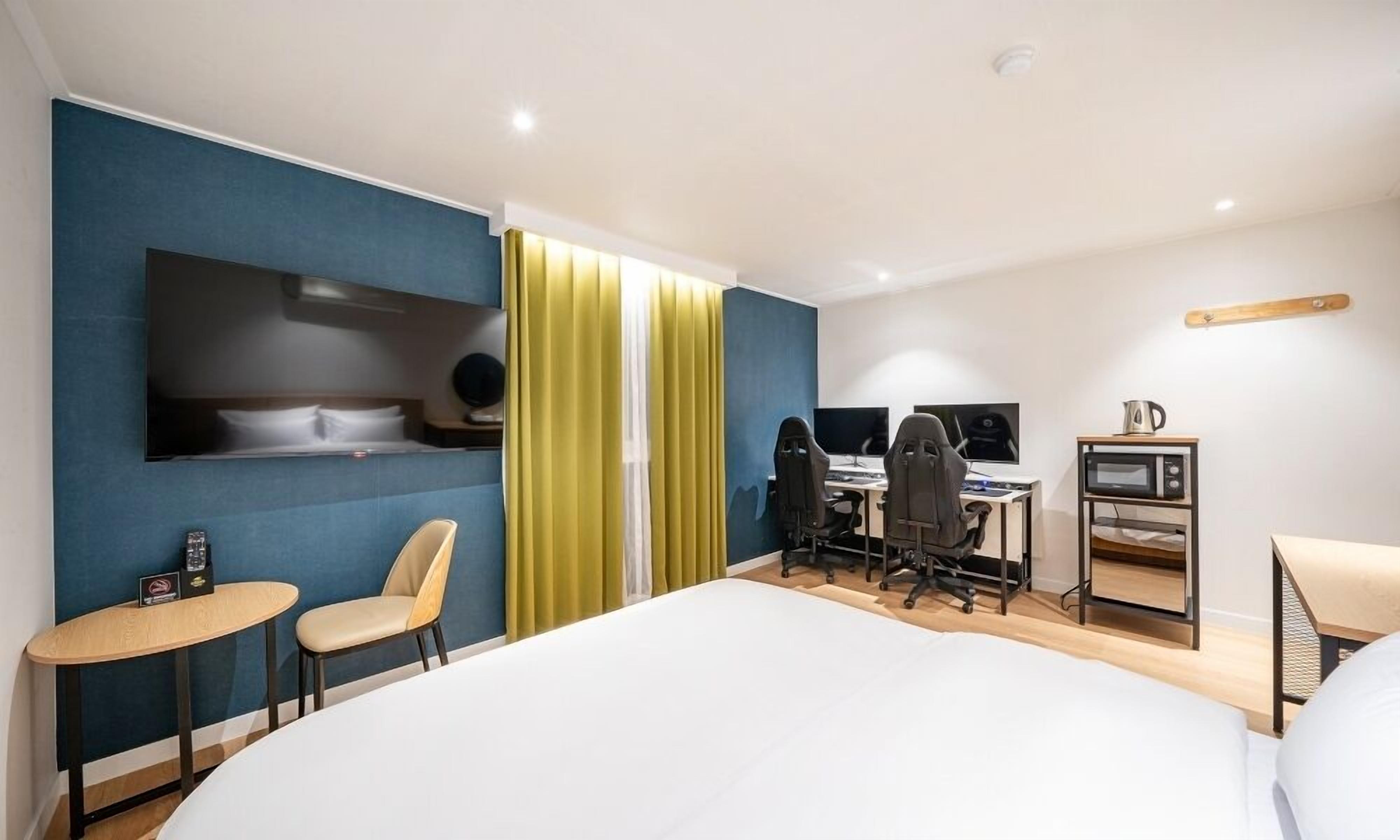 Habitación de hotel con una cama grande, un escritorio con dos monitores, una silla, una mesita y un televisor montado en la pared.