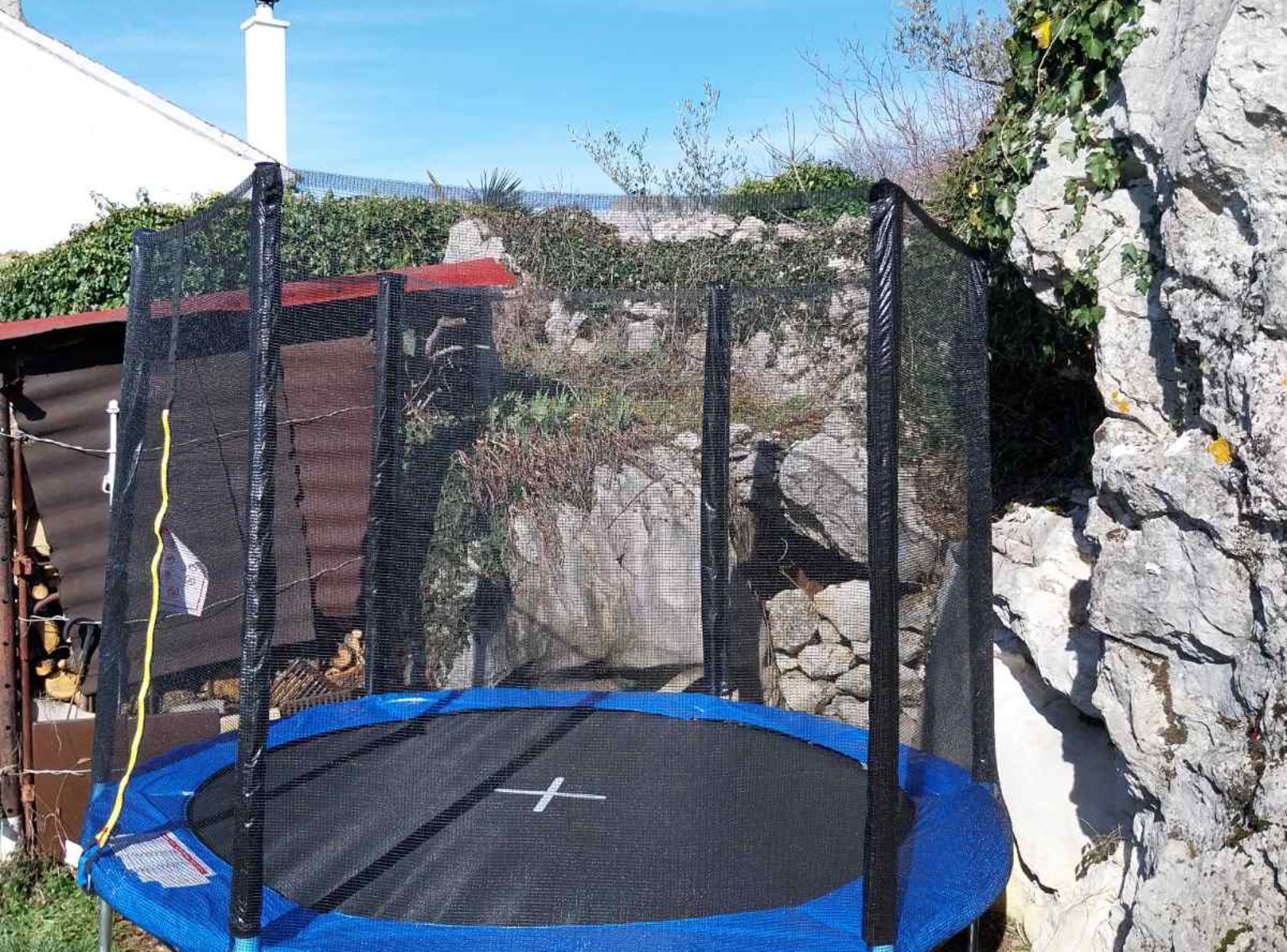 A trampoline enclosed by a safety net, set against a stone wall and a red-roofed building.