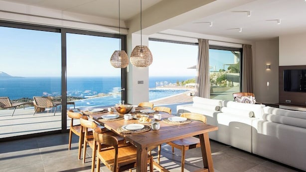 Una sala de estar moderna con un ventanal grande que da a una piscina y al océano, un comedor con muebles de madera y un sofá blanco.