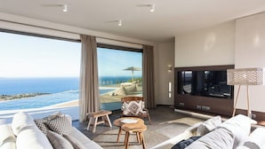 Una sala moderna con un televisor grande, un sofá, una mesita y un sillón. Hay un ventanal con vistas al océano y a la playa.