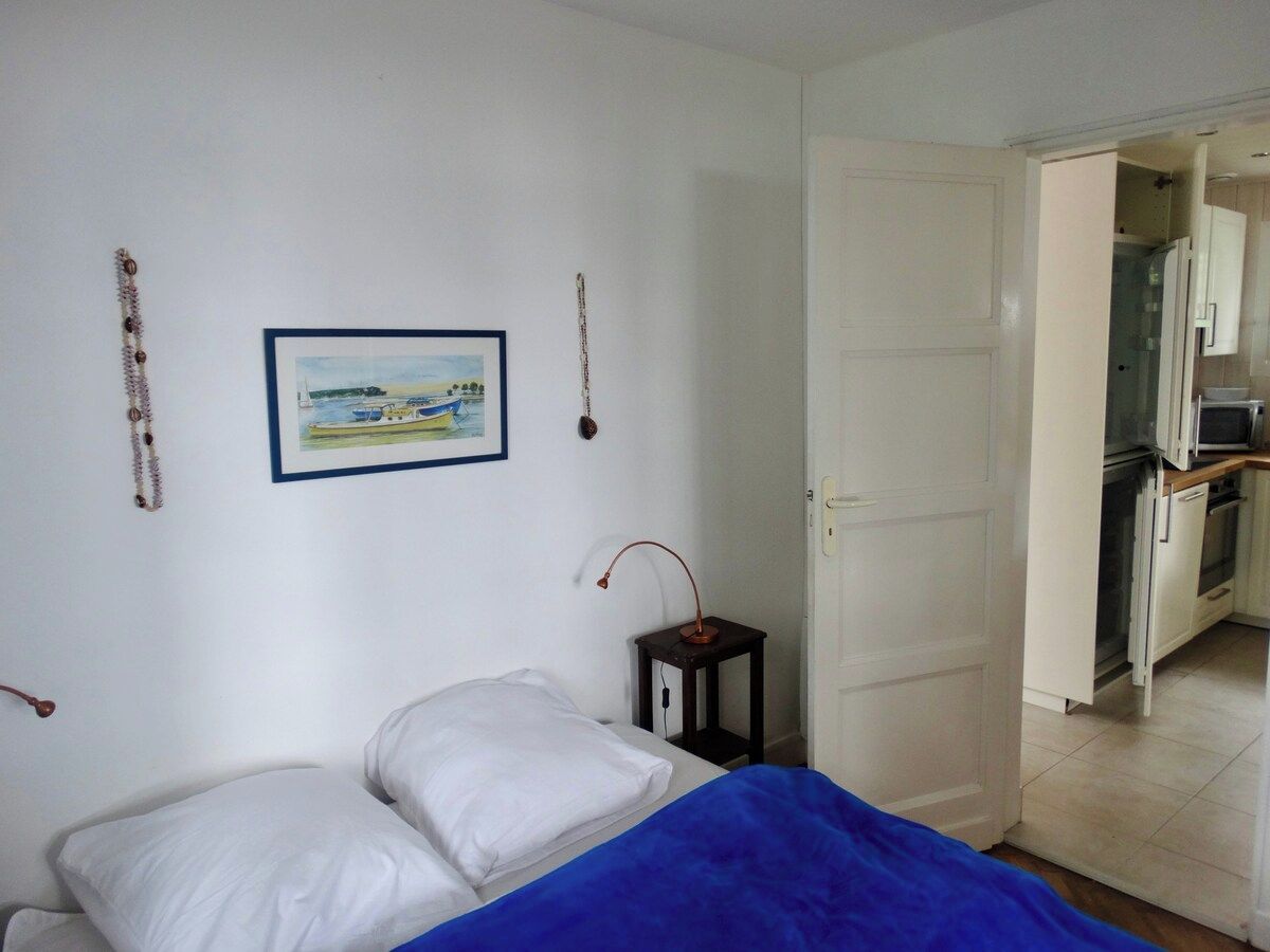 Une chambre à coucher avec un lit, une table de chevet, un tableau accroché au mur et une porte ouverte donnant sur un coin cuisine.