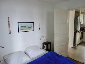 A bedroom with a bed, a nightstand, a picture on the wall, and an open door to a kitchen area.