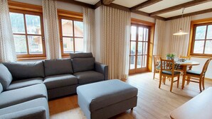 A cozy living room with a sectional sofa, a dining table with chairs, and large windows with curtains.