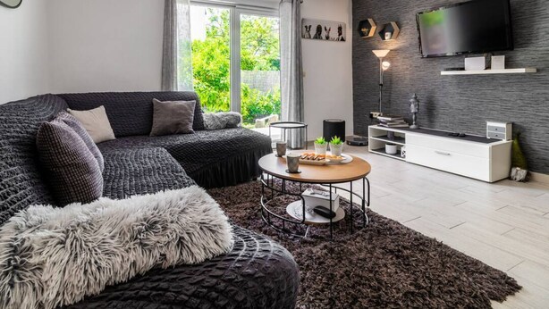 A modern living room with a dark grey sofa, a round coffee table, and a fluffy rug. A flat-screen TV is mounted on the wall, and there is a window with curtains. - Holiday Home Sunny Side (Kastela)