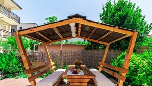 A wooden pergola with a table and benches, surrounded by greenery and a fence. - Holiday Home Sunny Side (Kastela)