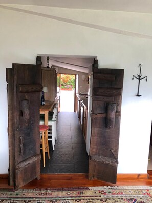 Un pasillo con puertas de madera rústica, un comedor con mesa y sillas, y vista a un jardín por una puerta abierta.