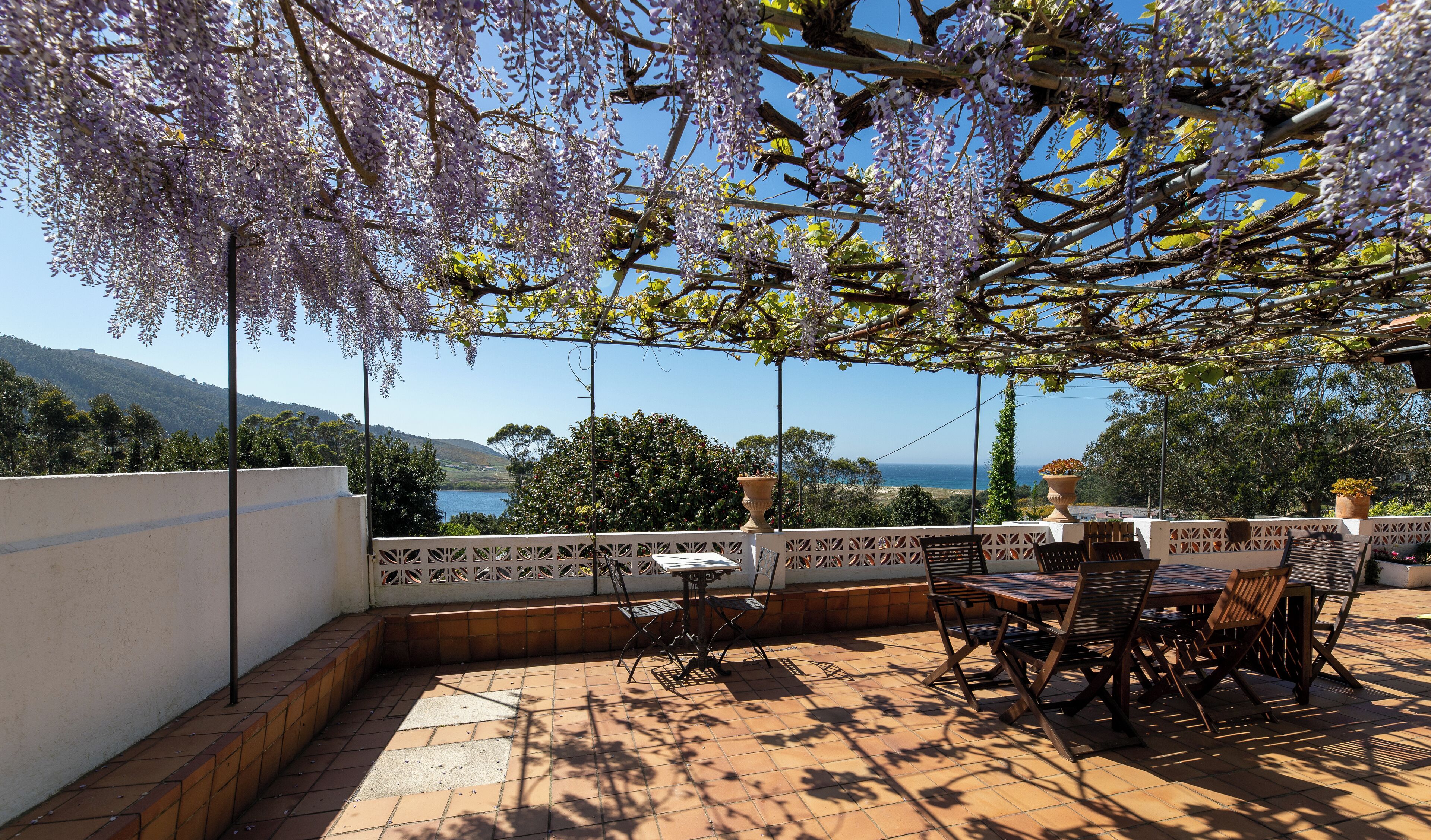 An outdoor patio with a table and chairs, a pergola with purple flowers, and a view of the sea and hills.