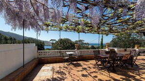 An outdoor patio with a table and chairs, a pergola with purple flowers, and a view of the sea and hills. - La Entretenida -  View and Sound of the Sea, Next to the Beach. Pet-Friendly. (Doniños Ferrol)