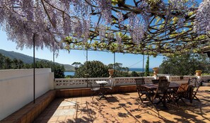 Eine Terrasse im Freien mit Tisch und Stühlen, eine Pergola mit violetten Blüten und Blick auf Meer und Hügel.