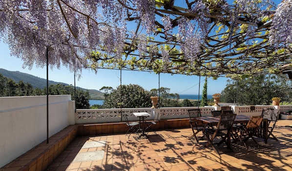 An outdoor patio with a table and chairs, a pergola with purple flowers, and a view of the sea and hills.