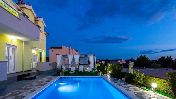 A swimming pool with a tiled edge, surrounded by a stone deck and a small garden area with a white canopy and lounge chairs. - Villa Ava (Sibenik)
