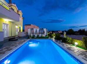 A swimming pool with a tiled edge, surrounded by a stone deck and a small garden area with a white canopy and lounge chairs. - Villa Ava (Sibenik)