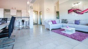 A modern living room with a white sofa, a coffee table, and a purple rug. There is a kitchen area with white cabinets and a dining table with chairs. - Villa Ava (Sibenik)
