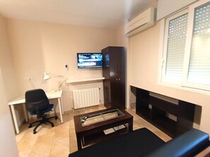 A small living room with a desk, a chair, a television, a fireplace, and a radiator. - Flamencos del Cerrado G-2 Small studio with mountain views (Málaga)