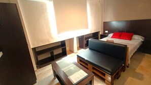 Une chambre d’hôtel de taille modeste, comprenant un lit, un petit bureau, une chaise, un ventilateur de plafond et une fenêtre avec des rideaux.