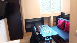 Un espace de vie compact comprenant un coin cuisine, une table à manger, un canapé agrémenté de coussins à motifs, un téléviseur fixé au mur et une fenêtre ornée d’une grille décorative.