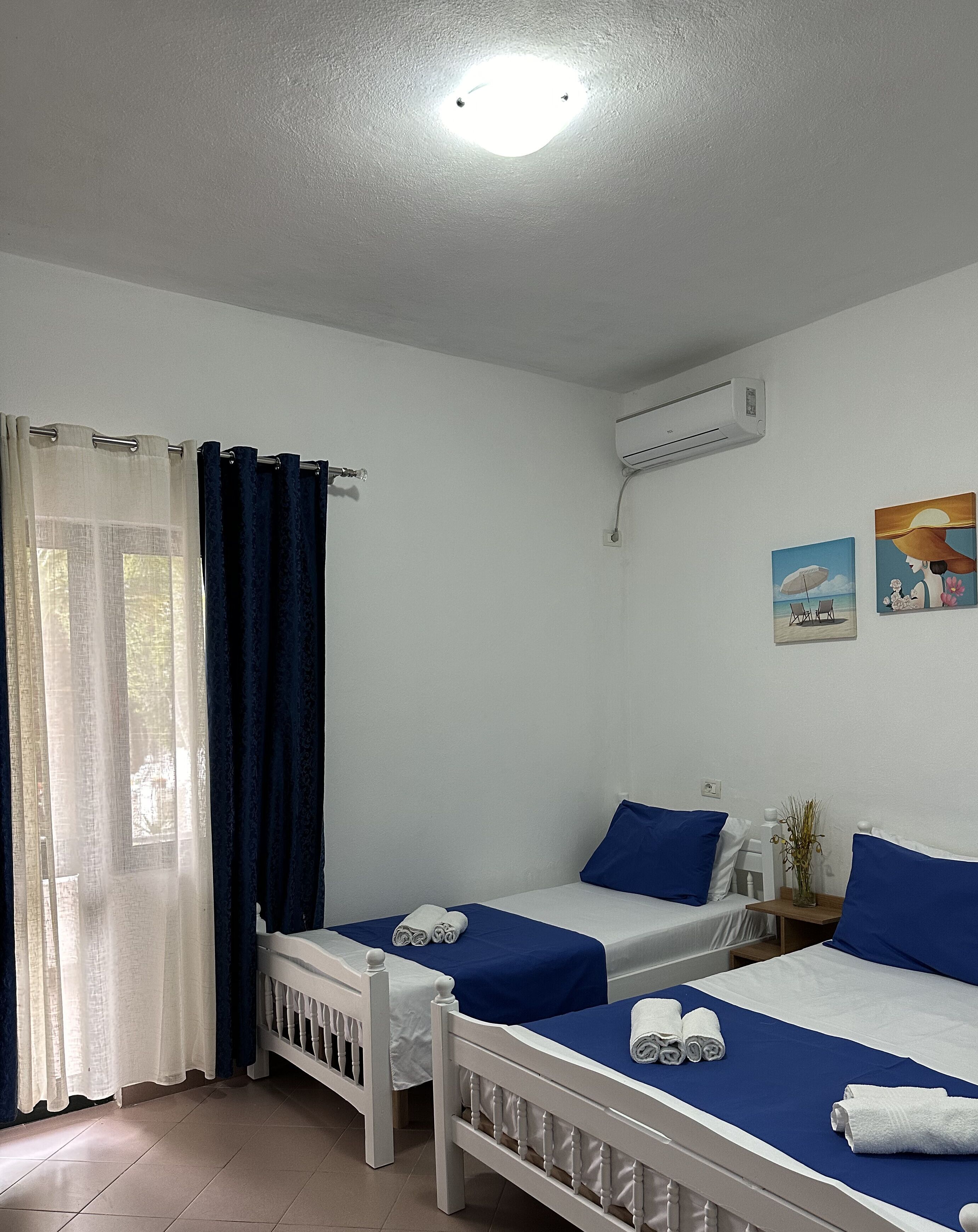 A room with two single beds, a window with curtains, an air conditioning unit, and a wall-mounted artwork.