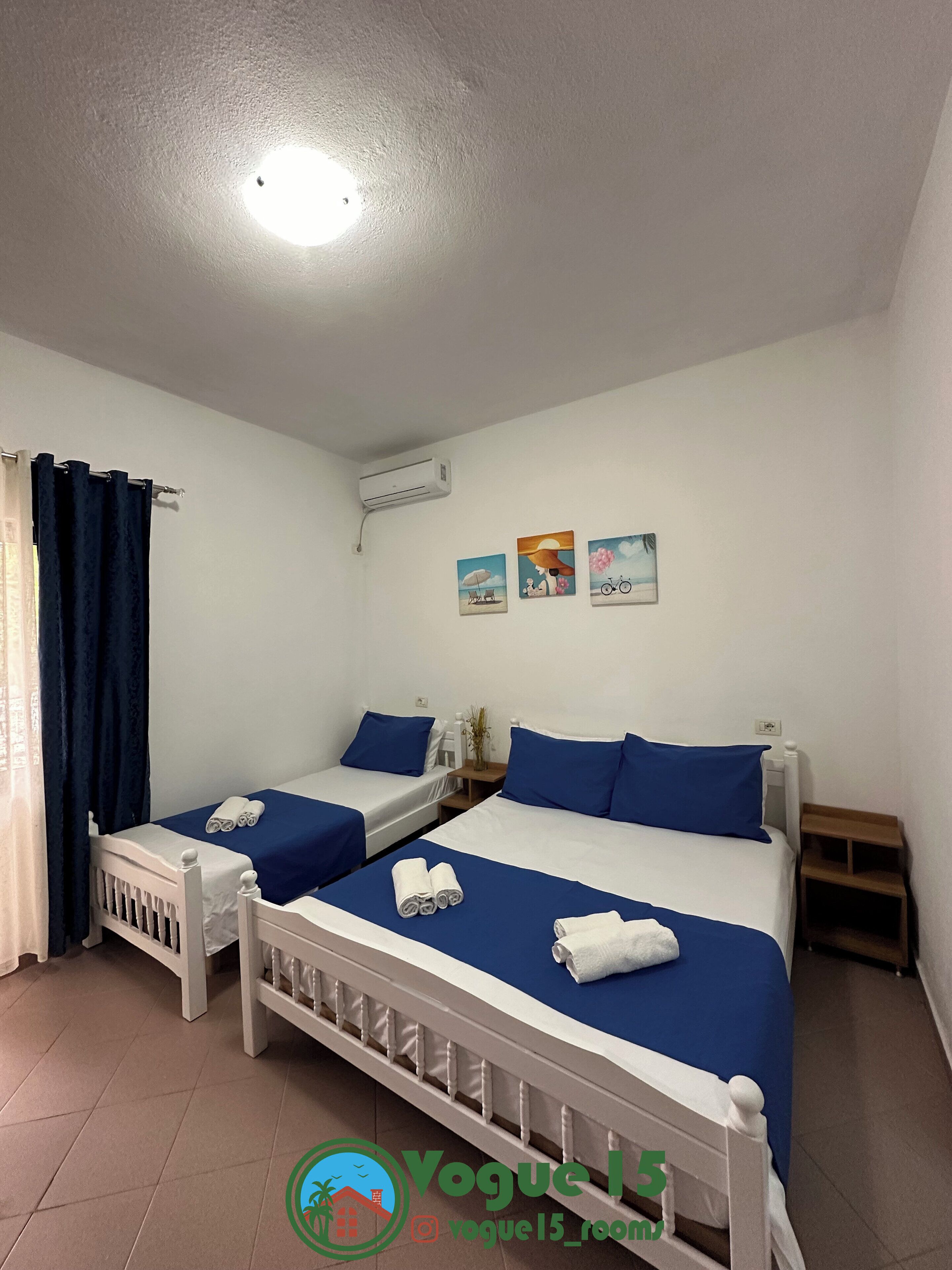 A room with two beds, each with blue bedding and white headboards, a small table with a vase, and a wall-mounted air conditioner.