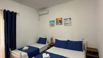 A room with two beds, each with blue bedding and white headboards, a small table with a vase, and a wall-mounted air conditioner.