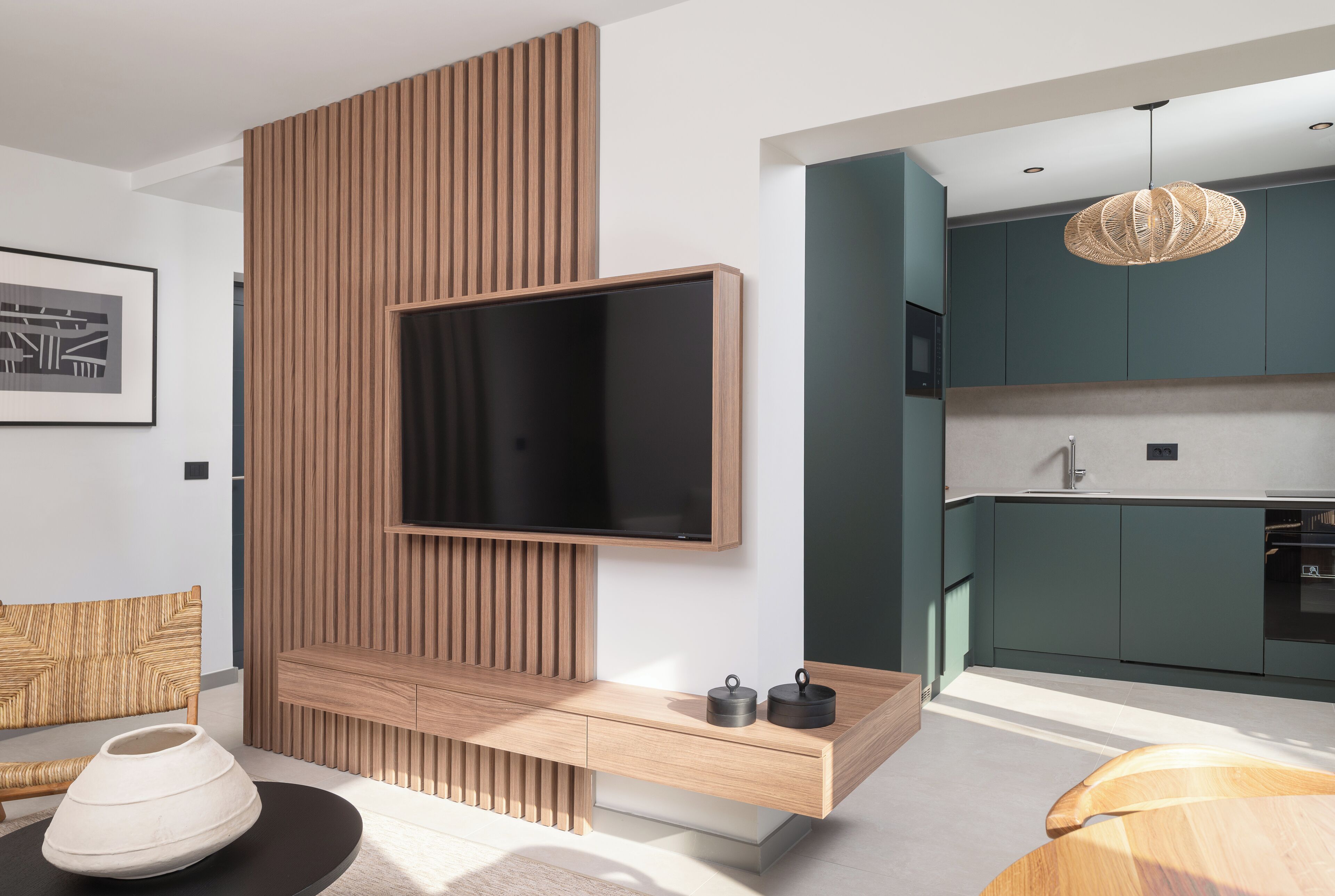 A modern living room with a TV mounted on a wooden panel wall, a sofa, a coffee table, and a kitchen area visible through an open door.