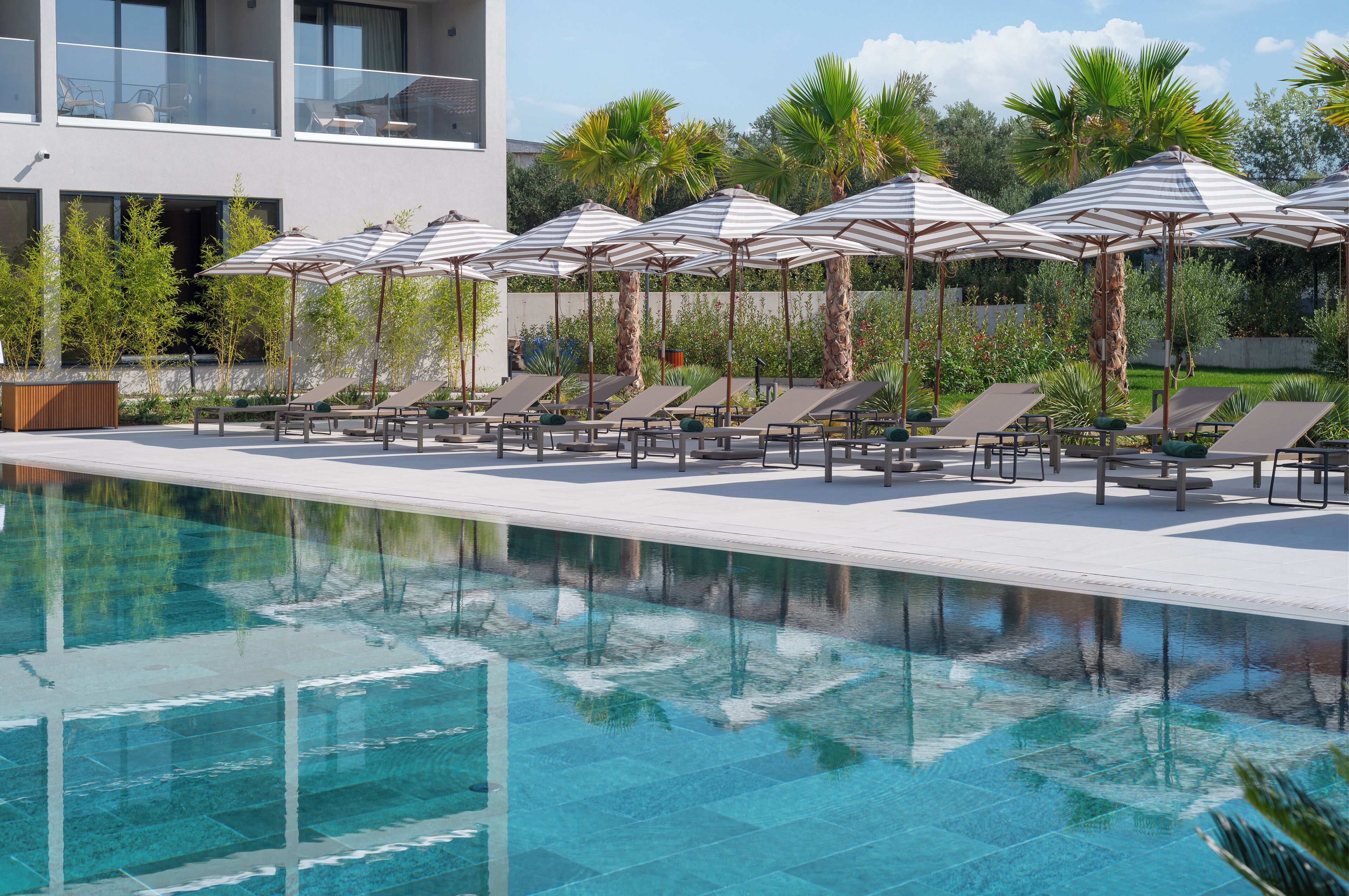Uma área de piscina com espreguiçadeiras e guarda-sóis, cercada por palmeiras e um edifício moderno.